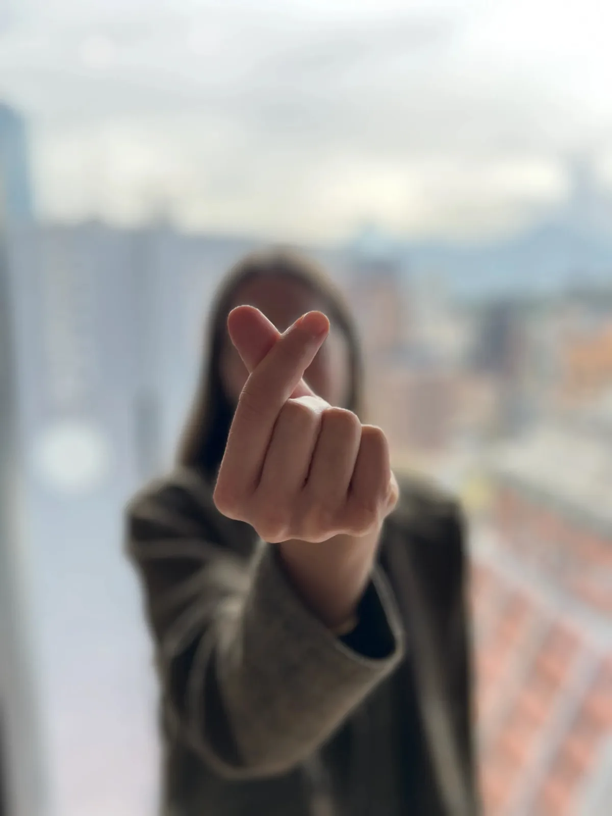 Persona haciendo un corazón con los dedos frente a una ventana con vista a la ciudad